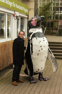 Michael Müller im Zoo vor dem mit einer Pinguinfigur vor dem Pavillion, in dem der Bau der Pinguinanlage erklärt wird